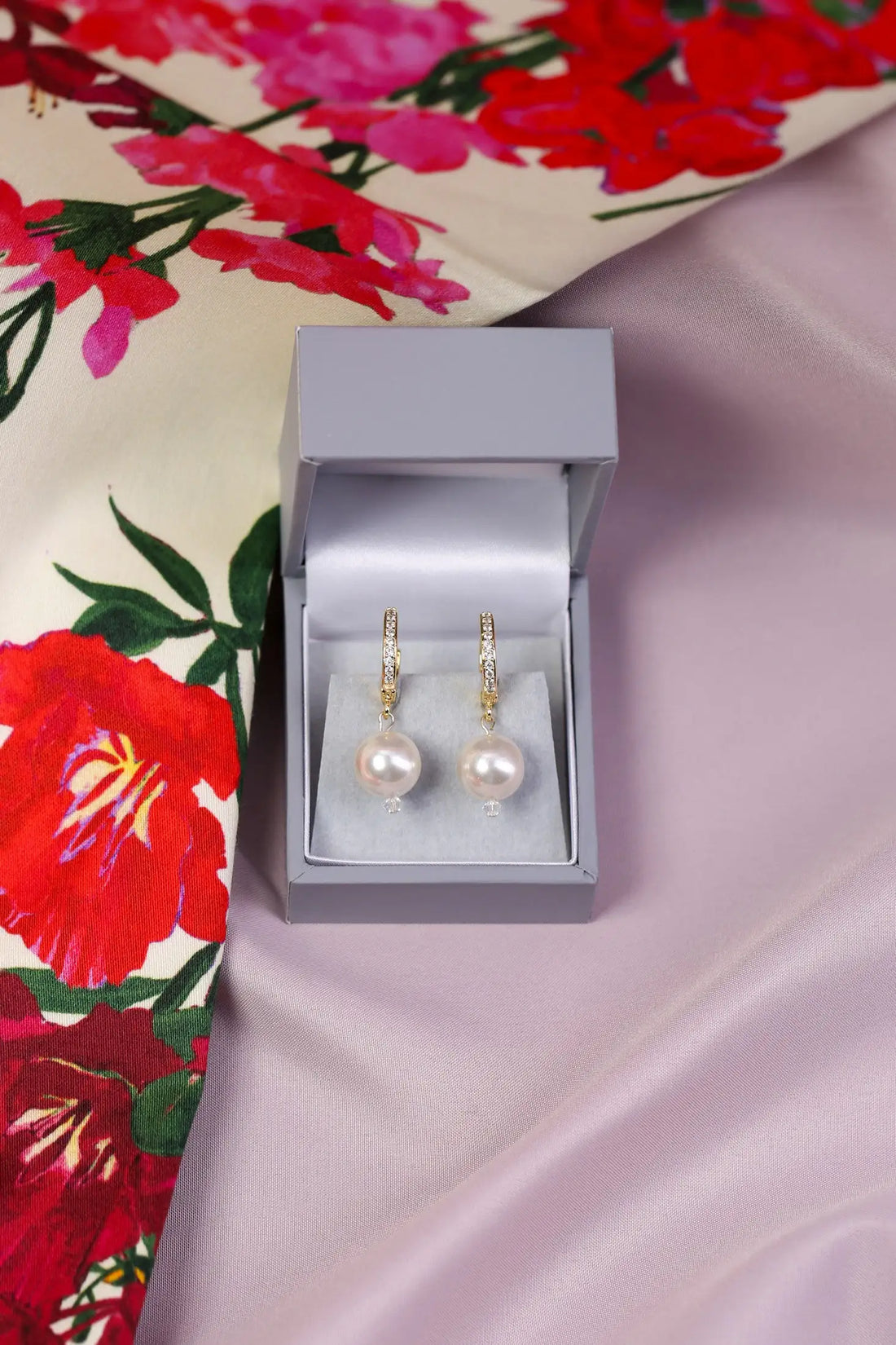 A pair of gold huggie earrings with pearls displayed in an open box, set against a floral patterned fabric background.