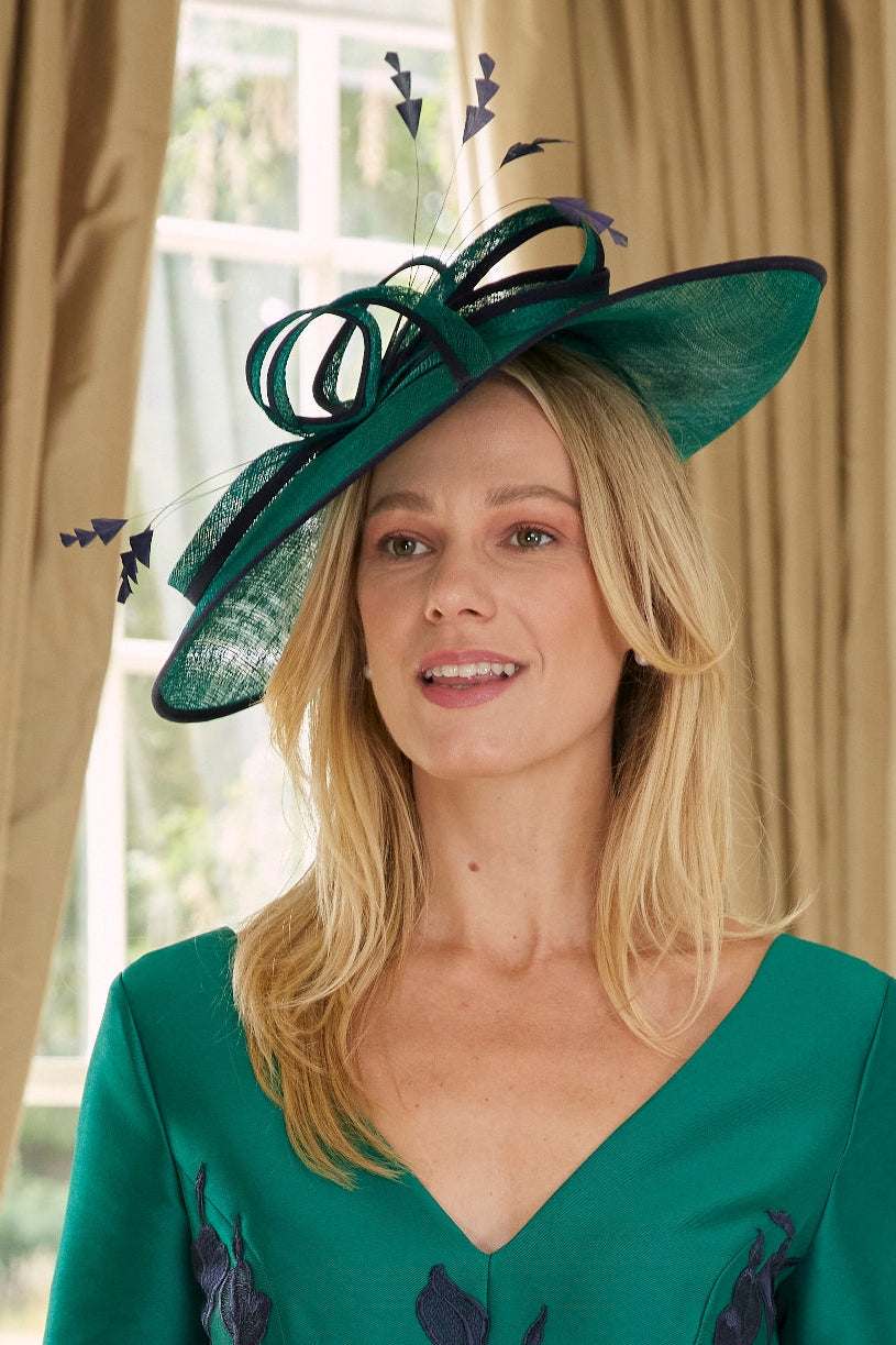 A woman wearing a green dress and matching green fascinator hat with dark navy floral embroidery and a decorative silver ring at the waist, posed indoors in front of beige curtains.