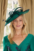 A woman wearing a green V-neck dress with dark navy floral embroidery, a matching dark navy belt with a jeweled circular buckle, and a matching green fascinator hat, posed in front of beige curtains.