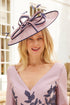 A woman wearing a lavender and navy dress with a matching wide-brimmed fascinator hat, standing outdoors in front of a white building.