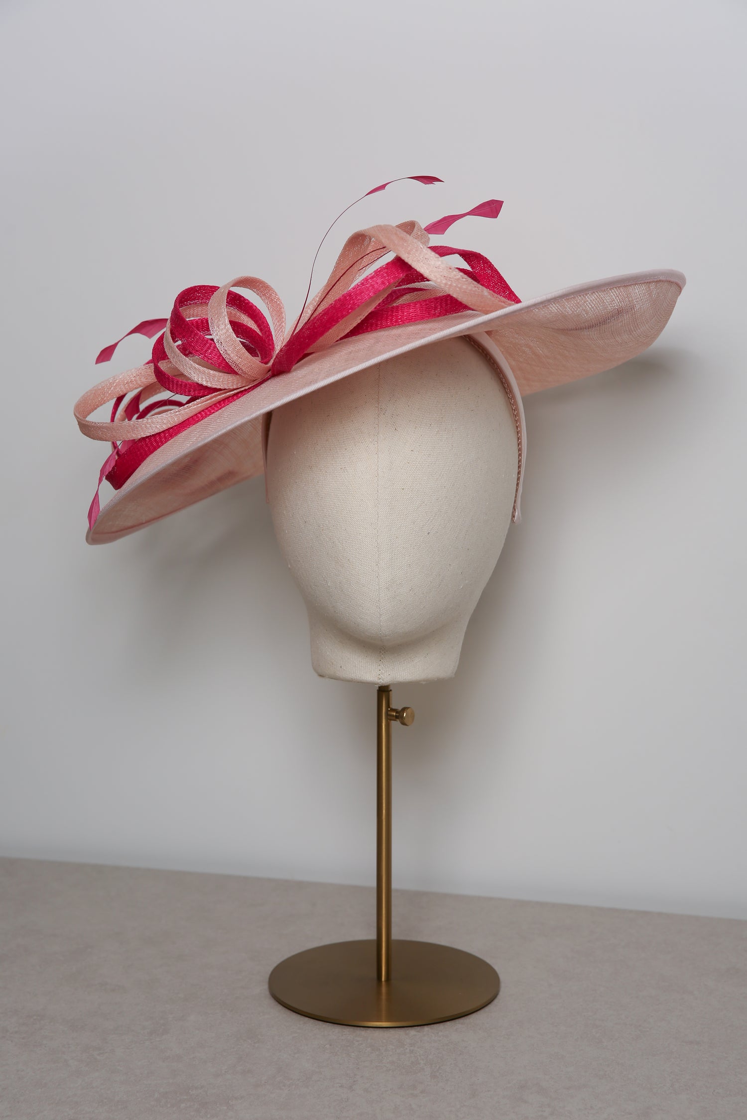 Pink and white decorative hat on a mannequin head with a gold stand against a plain background
