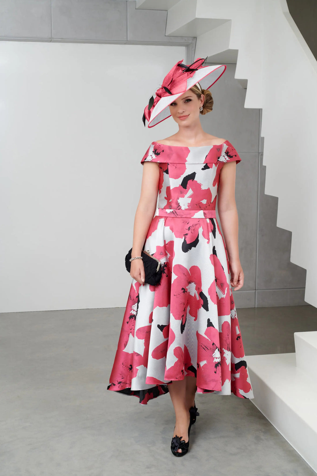 A model wearing an off-the-shoulder midi dress with a high-low hem, featuring a floral pattern in pink and white colours, paired with a hat and black shoes.