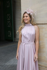 A woman wearing a pink sleeveless satin dress with a high neck and self-tie waistband, standing in front of a building. Styled with a matching fascinator.