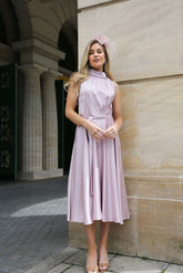 A woman wearing a pink sleeveless satin dress with a high neck and self-tie waistband, standing in front of a building.