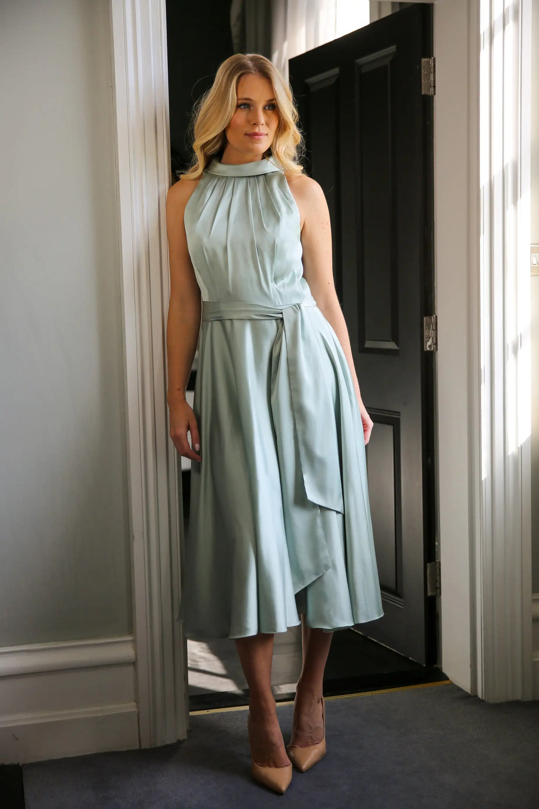 A woman wearing a sleeveless, high neck, seagrass colour satin midi dress with a self-tie waistband and pleating at the neckline.