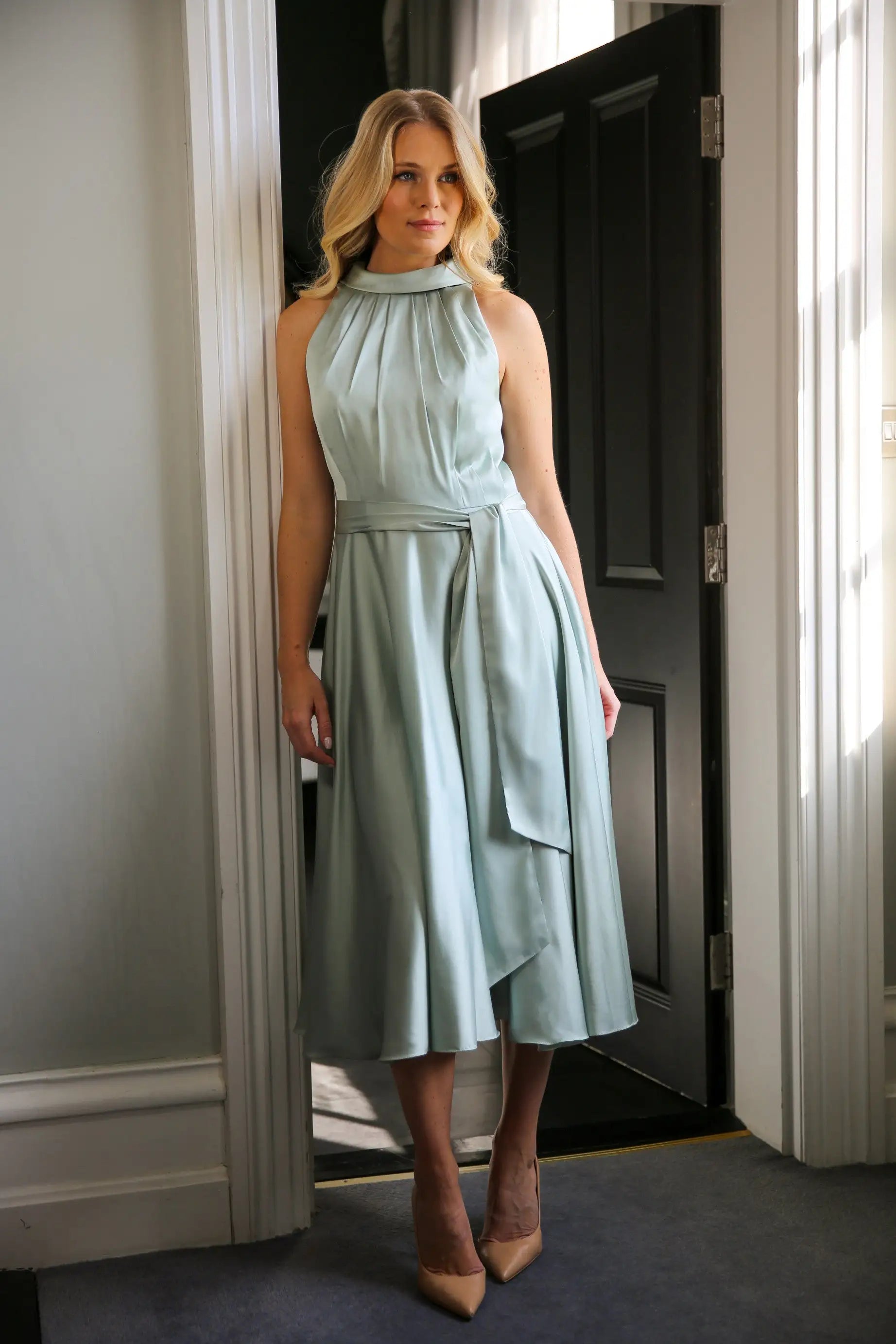 A woman wearing a sleeveless, high neck, seagrass colour satin midi dress with a self-tie waistband and pleating at the neckline.