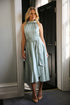 A woman wearing a sleeveless, high neck, seagrass colour satin midi dress with a self-tie waistband and pleating at the neckline.