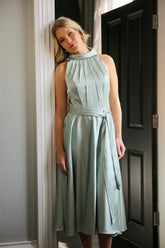 A woman wearing a sleeveless, high neck, seagrass colour satin midi dress with a self-tie waistband and pleating at the neckline.
