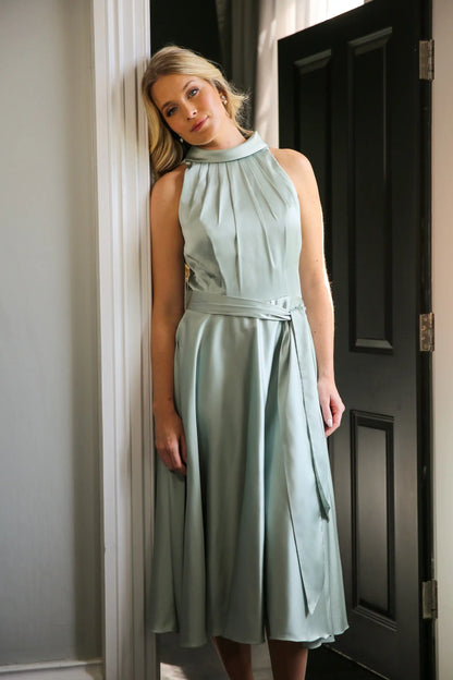 A woman wearing a sleeveless, high neck, seagrass colour satin midi dress with a self-tie waistband and pleating at the neckline.