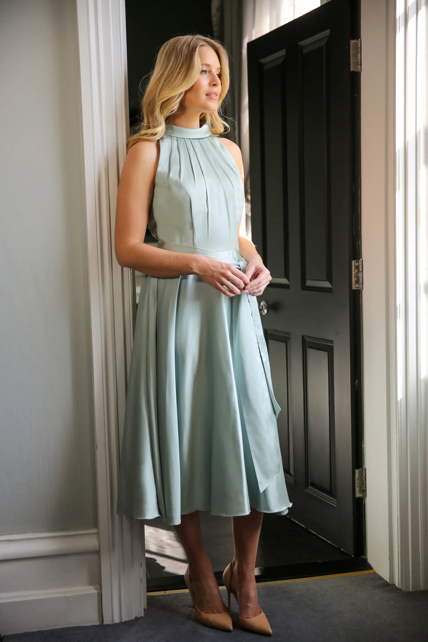 A woman wearing a sleeveless, high neck, seagrass colour satin midi dress with a self-tie waistband and pleating at the neckline.