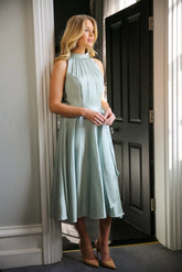 A woman wearing a sleeveless, high neck, seagrass colour satin midi dress with a self-tie waistband and pleating at the neckline.