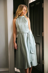 A woman wearing a sleeveless, high neck, seagrass colour satin midi dress with a self-tie waistband and pleating at the neckline.