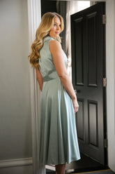 A woman wearing a sleeveless, high neck, seagrass colour satin midi dress with a self-tie waistband and pleating at the neckline.