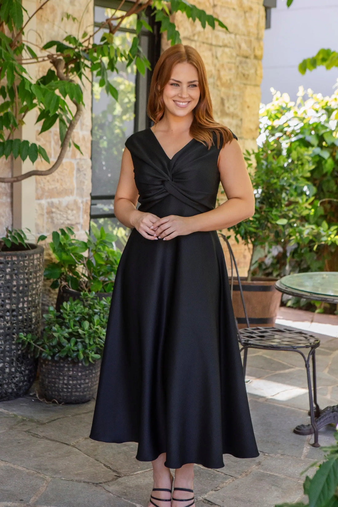 Woman in a black dress standing outdoors with plants and a stone wall in the background