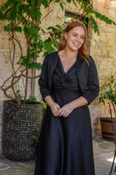 Woman in a black dress standing outdoors with plants and stone wall in the background