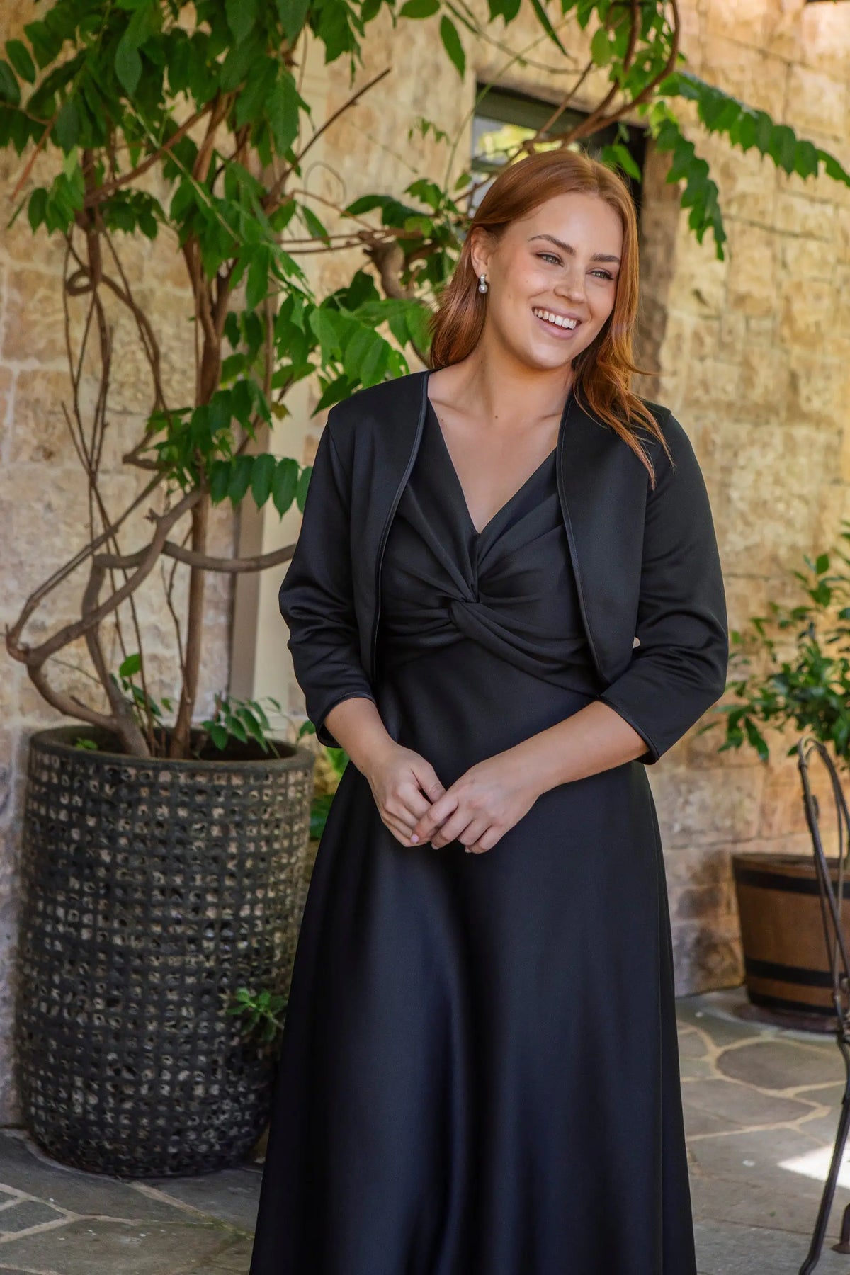 Woman in a black dress standing outdoors with plants and stone wall in the background