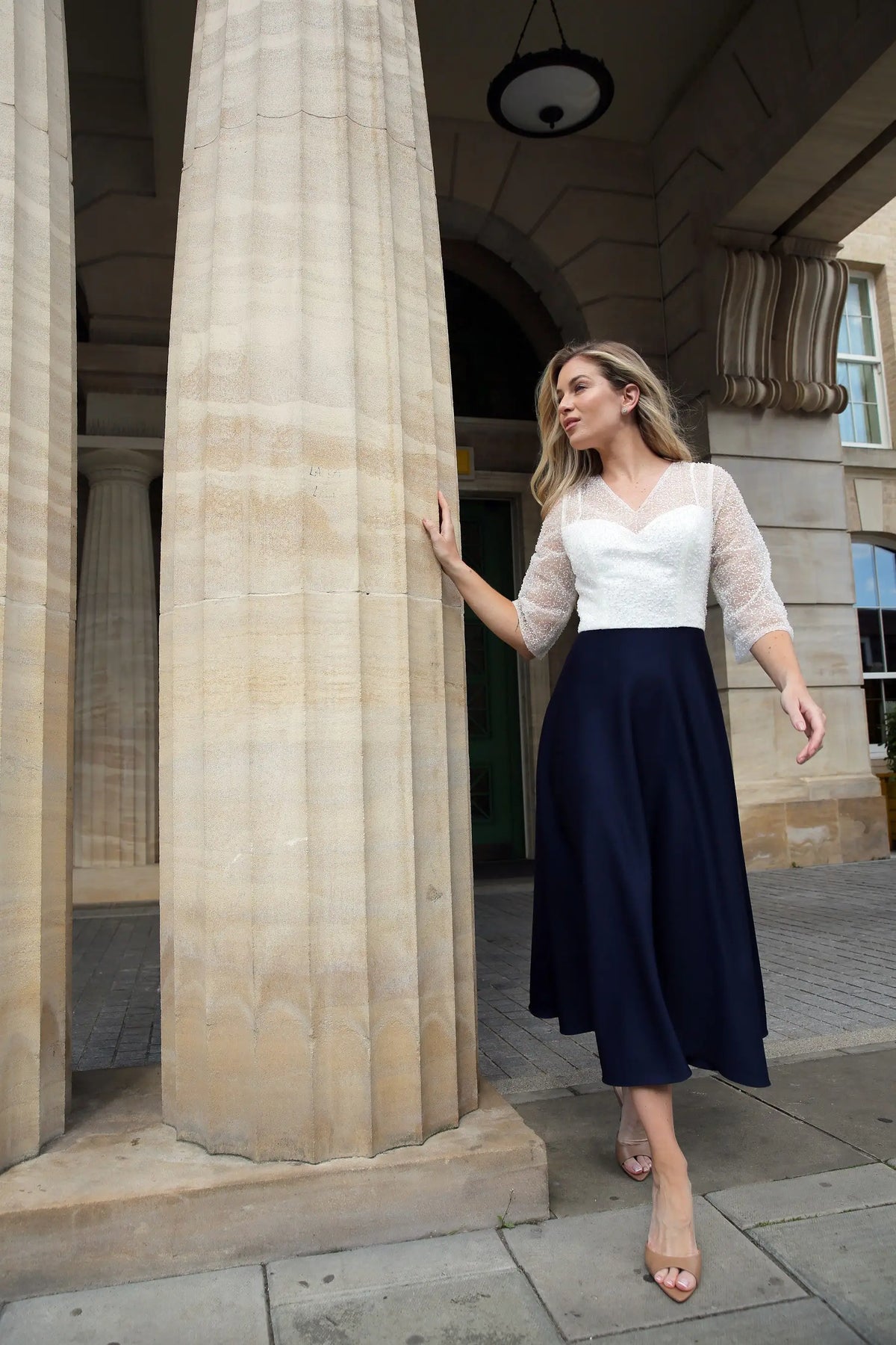 A woman standing outdoors wearing a dark navy, fully beaded bodice and sleeves, midi length dress with a sheer neckline and sleeves, and a back zip fastener.