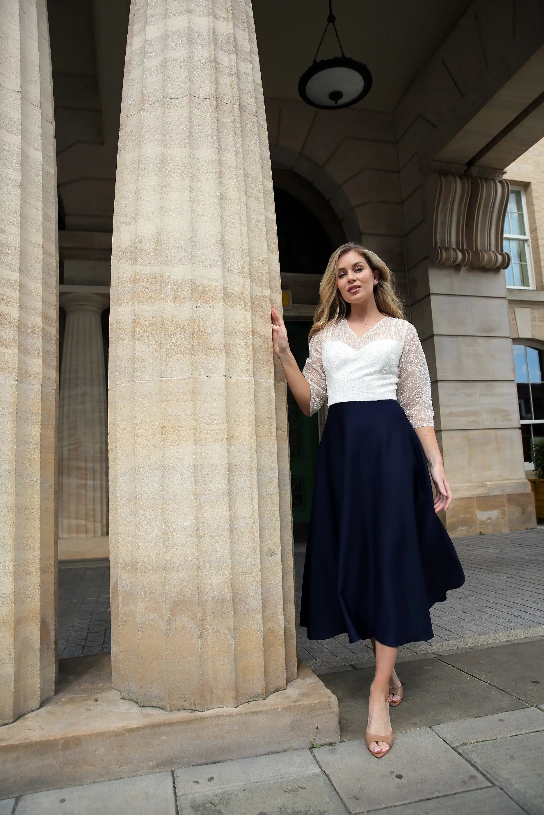 A woman standing outdoors wearing a dark navy, fully beaded bodice and sleeves, midi length dress with a sheer neckline and sleeves, and a back zip fastener.