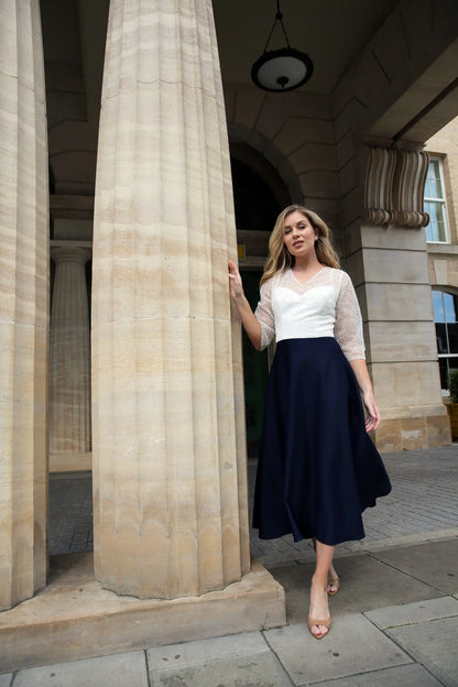 A woman standing outdoors wearing a dark navy, fully beaded bodice and sleeves, midi length dress with a sheer neckline and sleeves, and a back zip fastener.