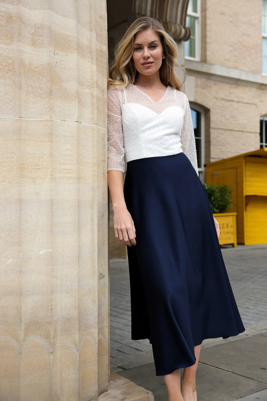 A woman standing outdoors wearing a dark navy, fully beaded bodice and sleeves, midi length dress with a sheer neckline and sleeves, and a back zip fastener.