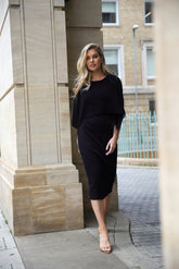 A woman standing in an elegant pose wearing a knee-length sleeveless black dress with a chiffon cape overlayer.