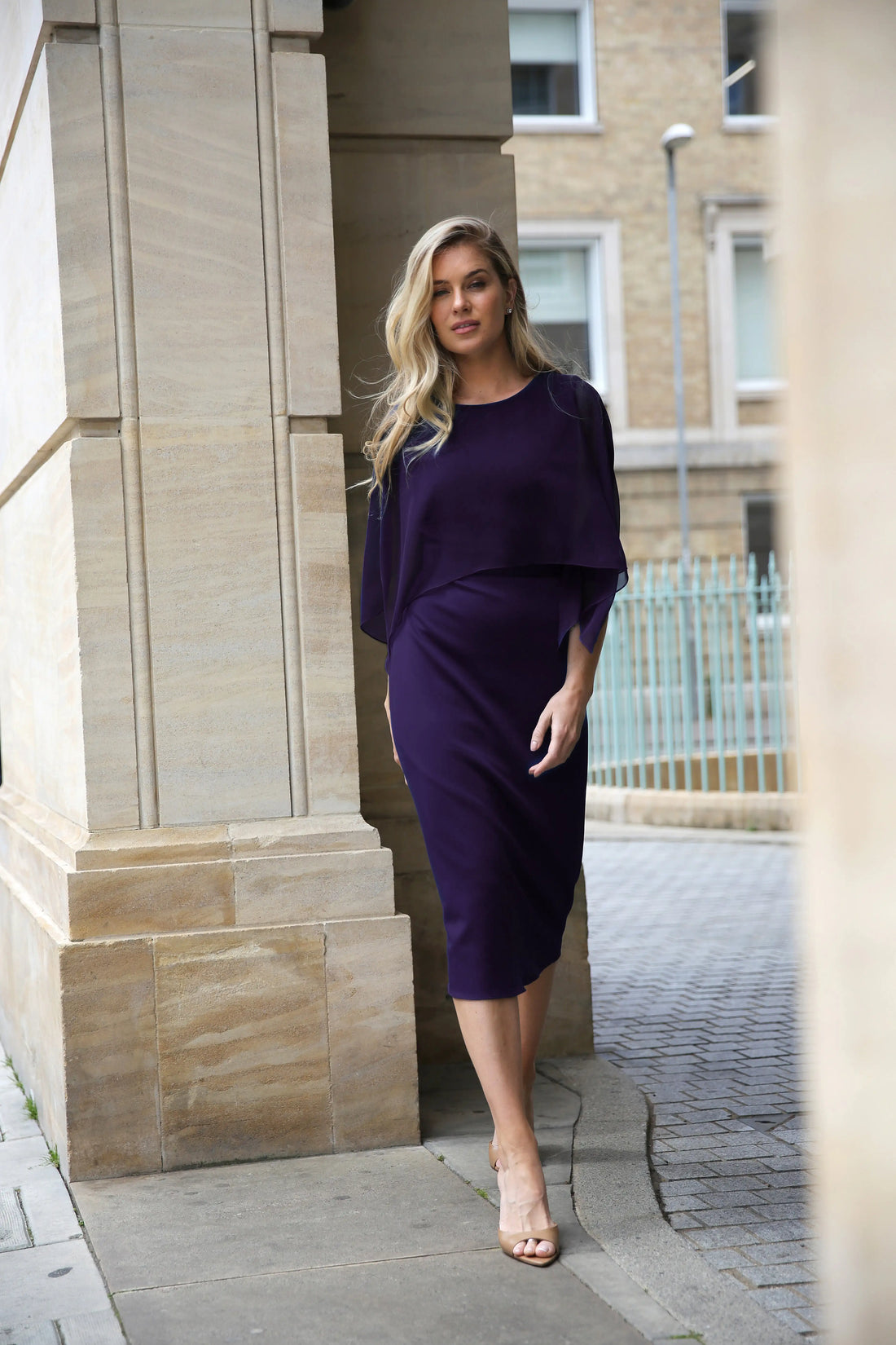 A woman standing in an elegant pose wearing a knee-length sleeveless dress with a chiffon cape overlayer.