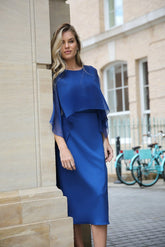A woman in an oxford blue jersey dress with a chiffon cape overlayer, standing on a street.