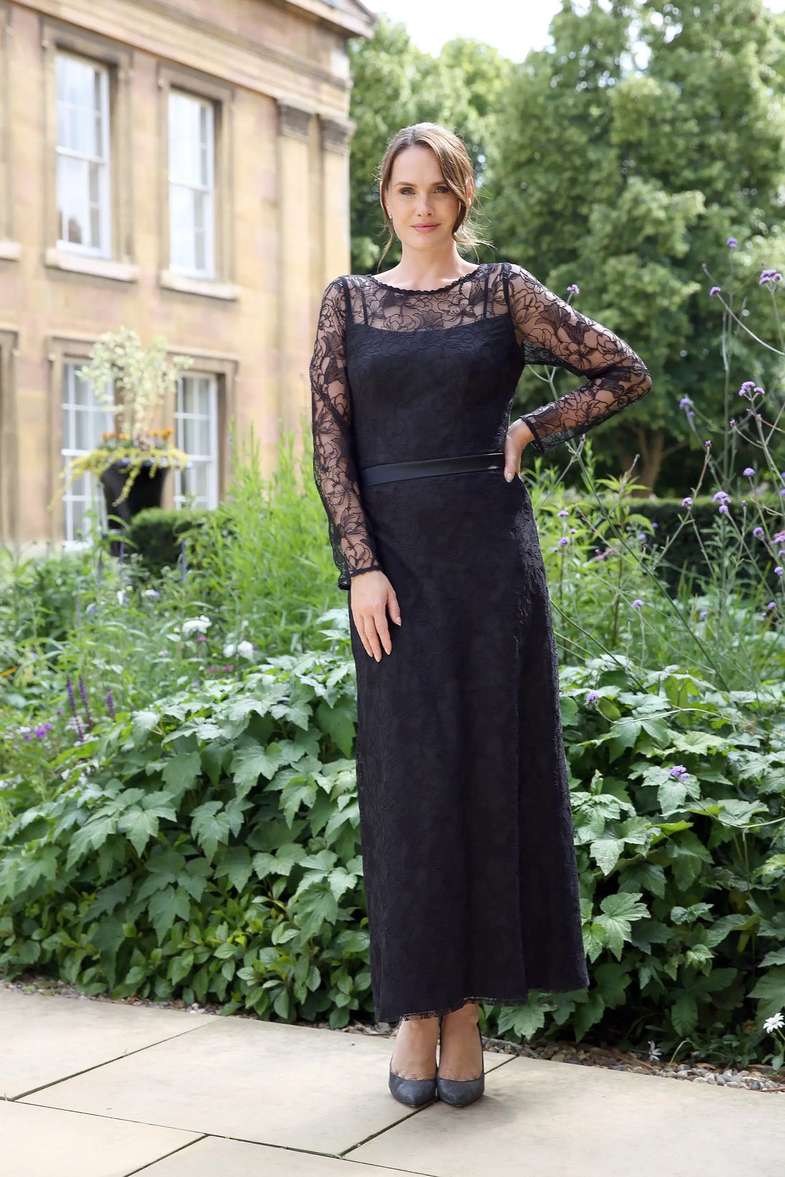 A woman wearing a black ankle-length evening dress with long sleeves and textured lace detailing.