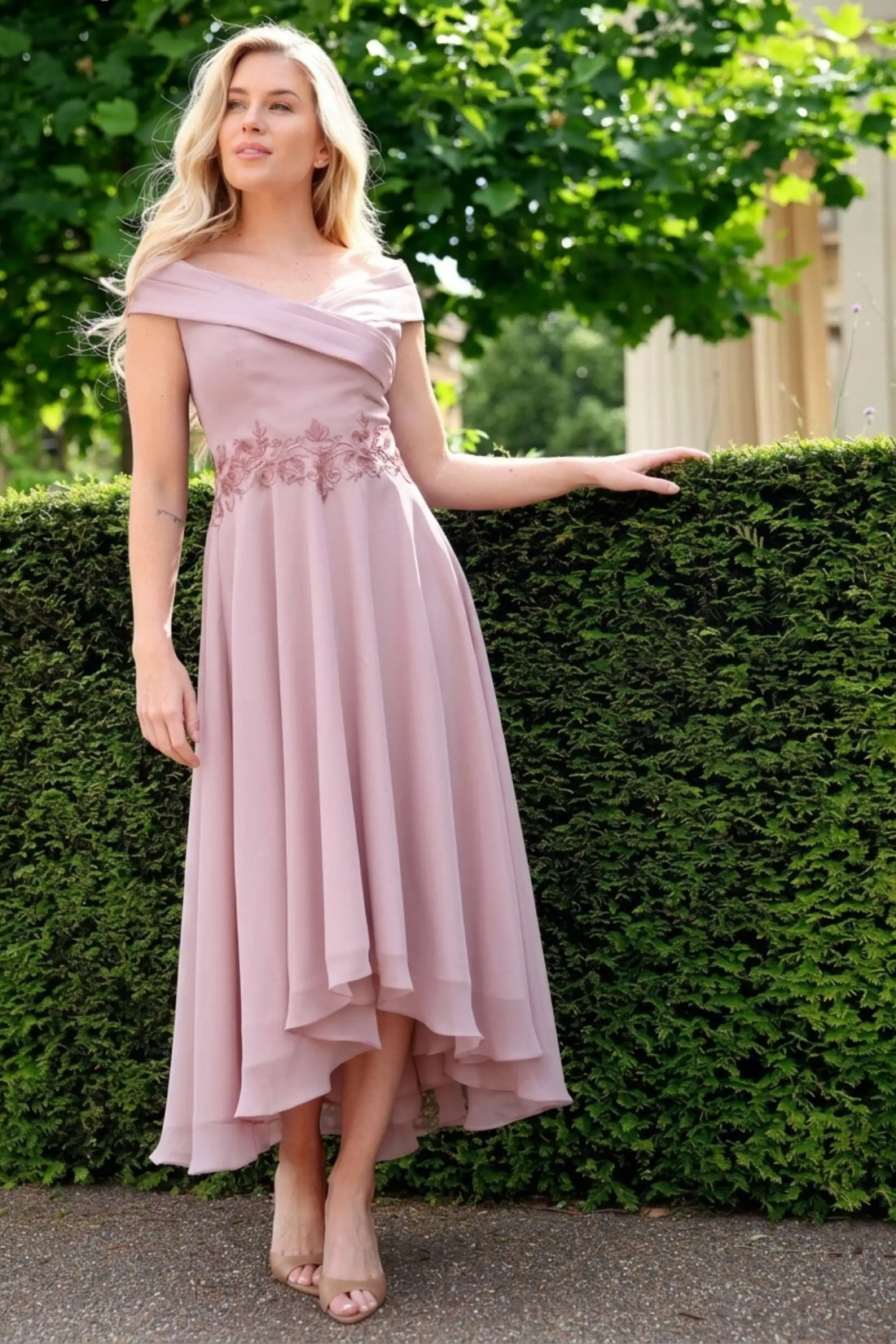 Woman in a pink off-shoulder dress standing against a green hedge.