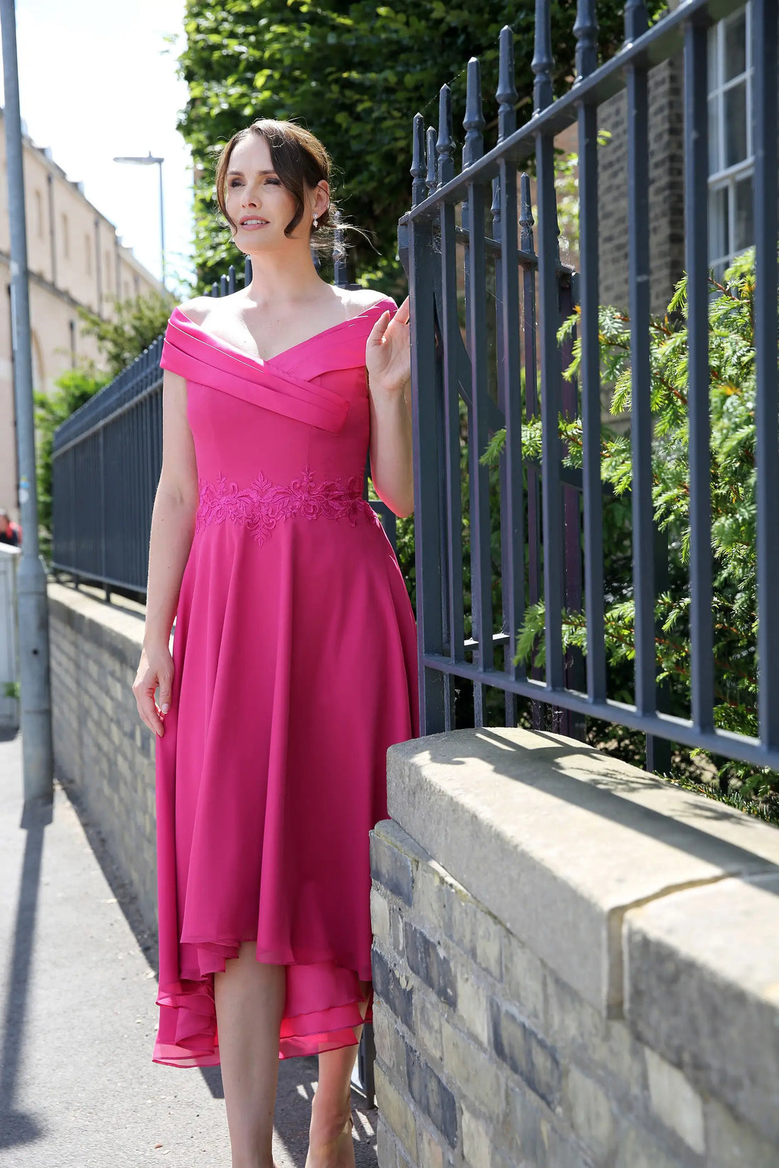 A woman standing by a black fence wearing a geranium pink, Bardot neckline, flowing chiffon high-low hem dress with embroidery on the waistline.