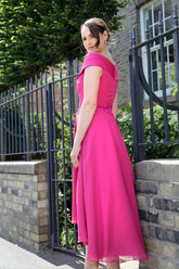 A woman standing by a black fence wearing a geranium pink, Bardot neckline, flowing chiffon high-low hem dress with embroidery on the waistline.