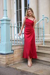 A woman in a red asymmetric ankle-length jersey dress with a draped bow detail over one shoulder, sleeveless with a back zip fastening.