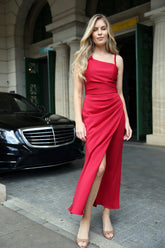 A woman in a red asymmetric ankle-length jersey dress with a draped bow detail over one shoulder, sleeveless with a back zip fastening.