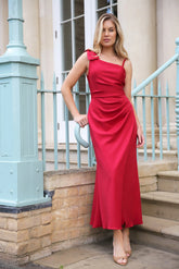 A woman in a red asymmetric ankle-length jersey dress with a draped bow detail over one shoulder, sleeveless with a back zip fastening.