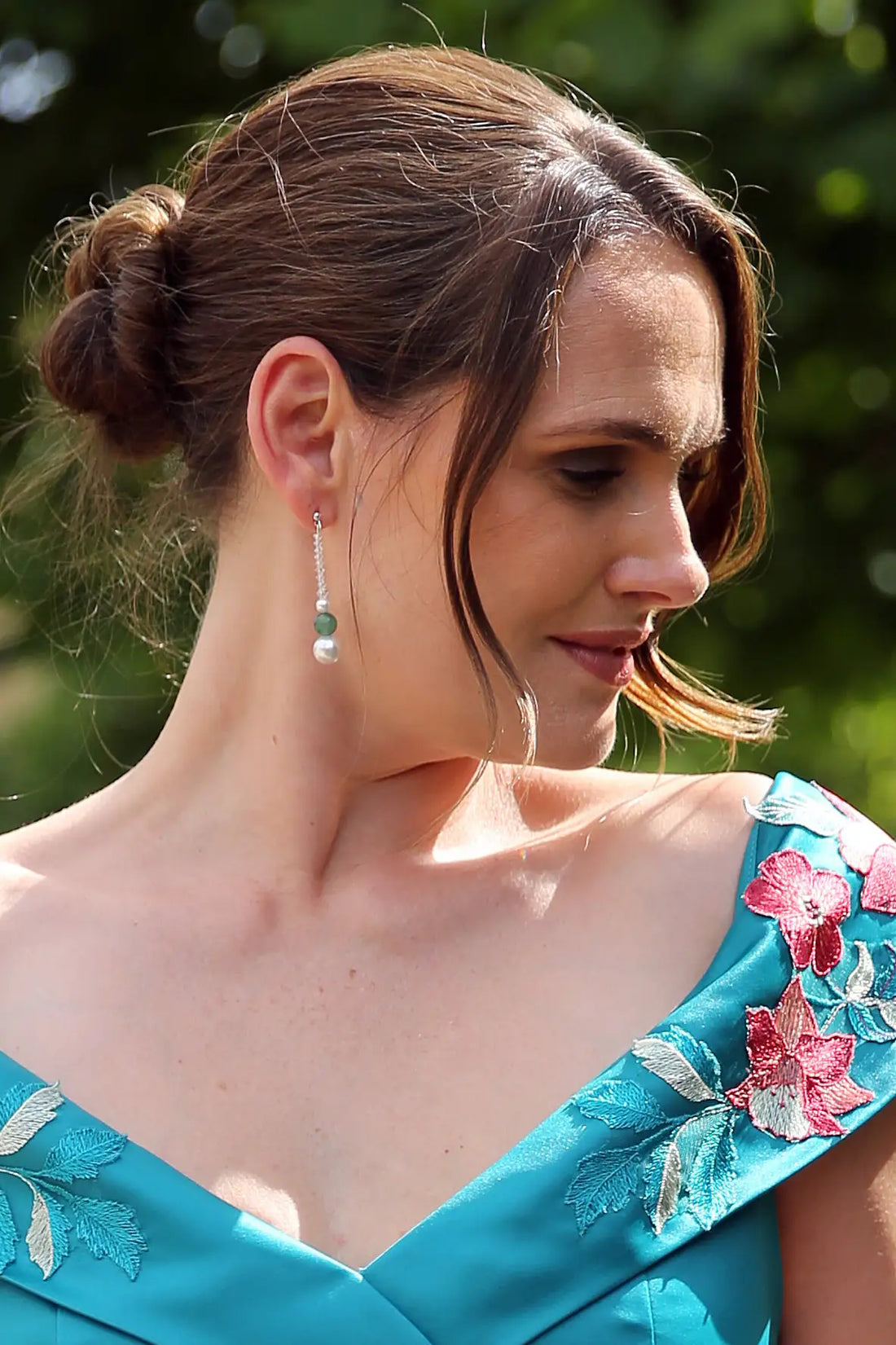 A person wearing sage green aventurine pearl drop earrings with a round pearl and a green bead, dressed in a blue garment with floral patterns.