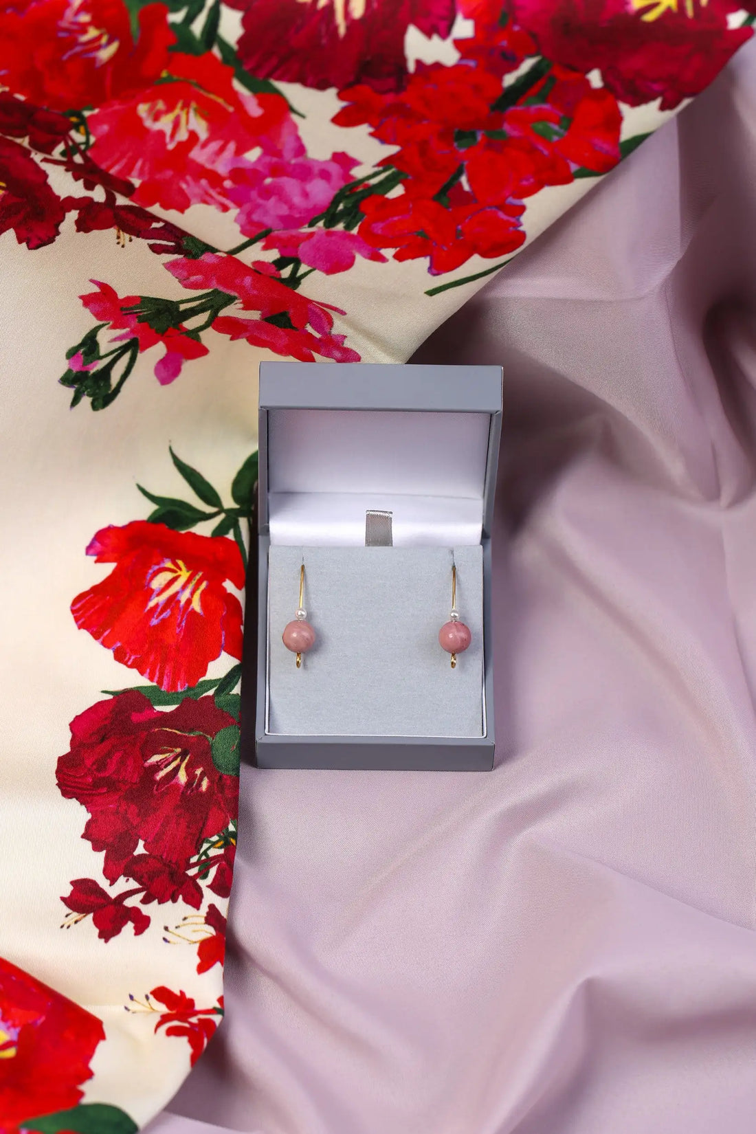 A pair of gold-coloured earrings with round cream pearls and mauve rhodonite beads, presented in a box with a floral patterned fabric background.