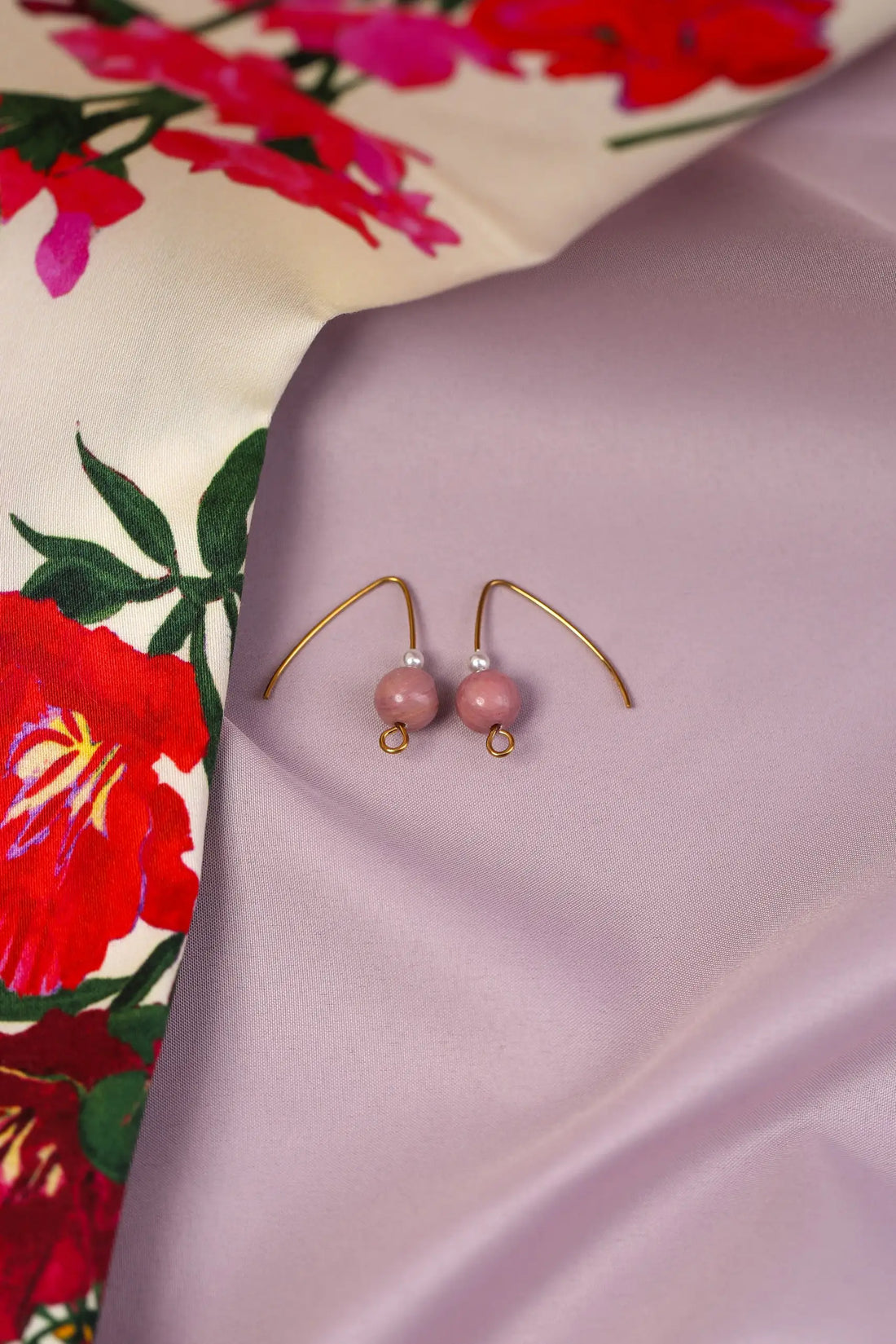 A pair of gold-coloured earrings with round cream pearls and mauve rhodonite beads, presented in a box with a floral patterned fabric background.