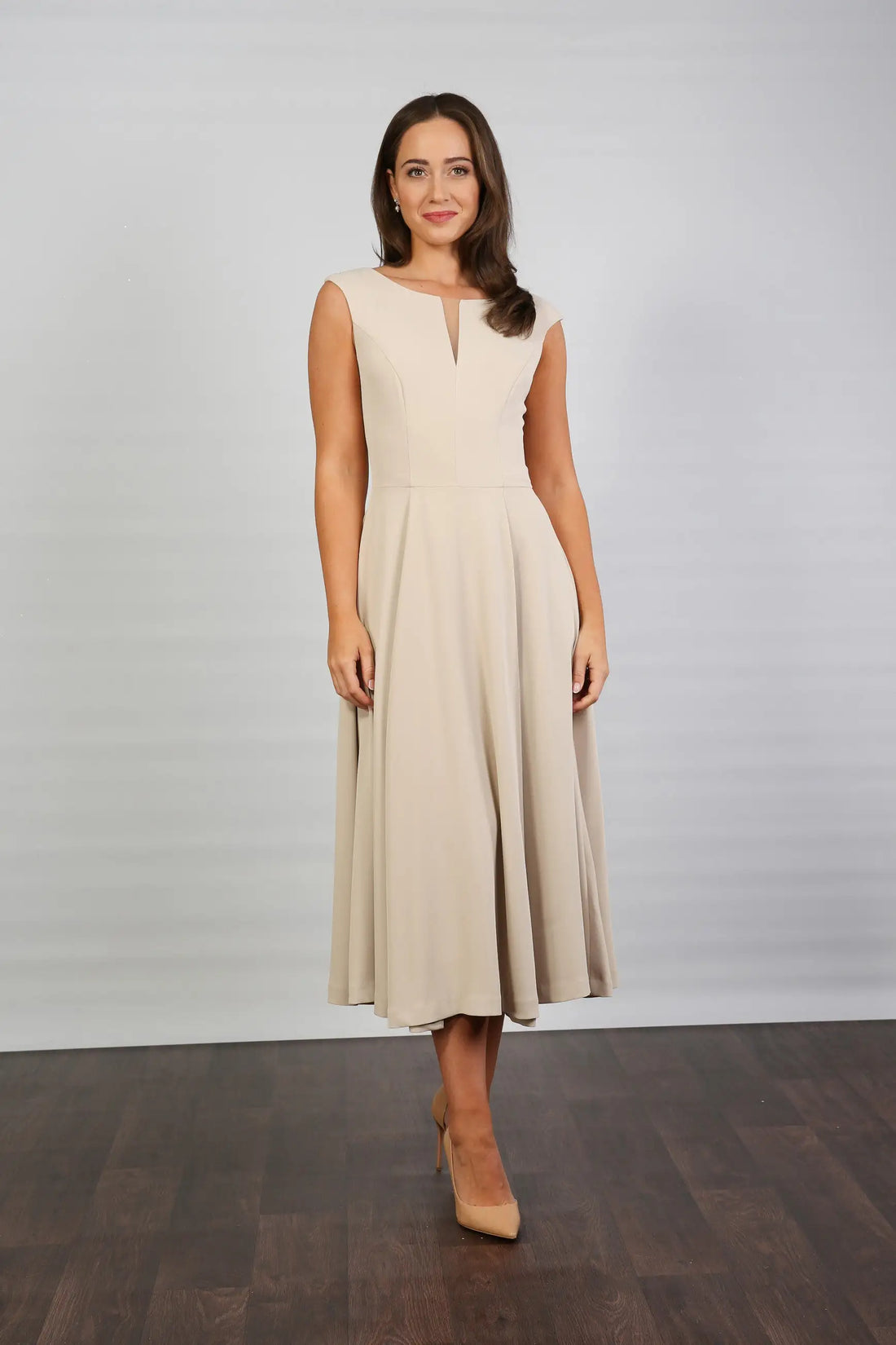 A woman standing in a semi-formal, sleeveless, midi length beige dress with a round neckline and a full skirt.
