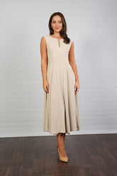 A woman standing in a semi-formal, sleeveless, midi length beige dress with a round neckline and a full skirt.