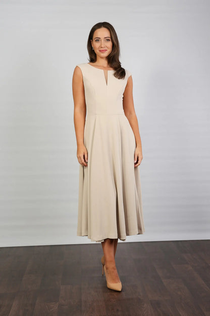 A woman standing in a semi-formal, sleeveless, midi length beige dress with a round neckline and a full skirt.