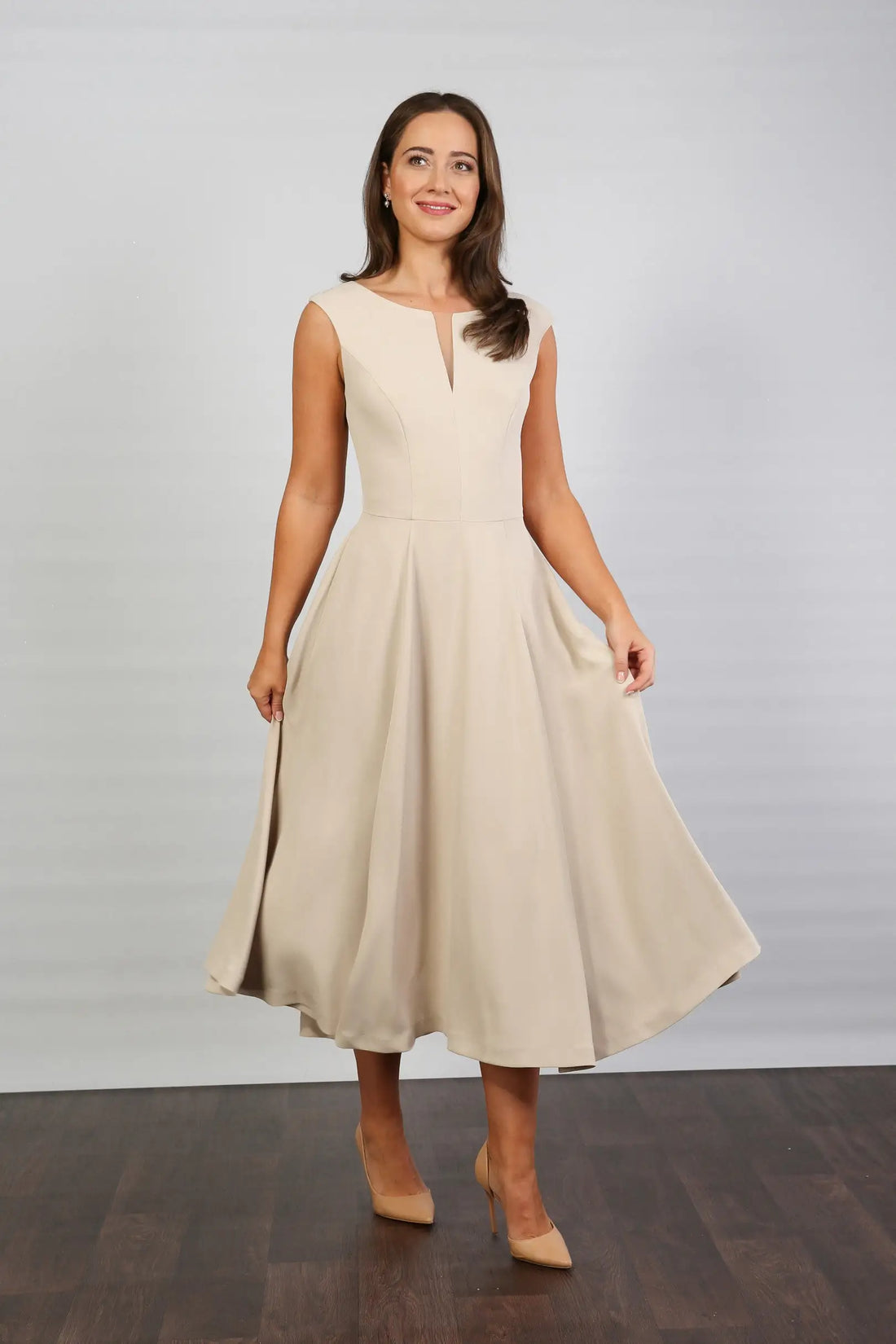 A woman standing in a semi-formal, sleeveless, midi length beige dress with a round neckline and a full skirt.