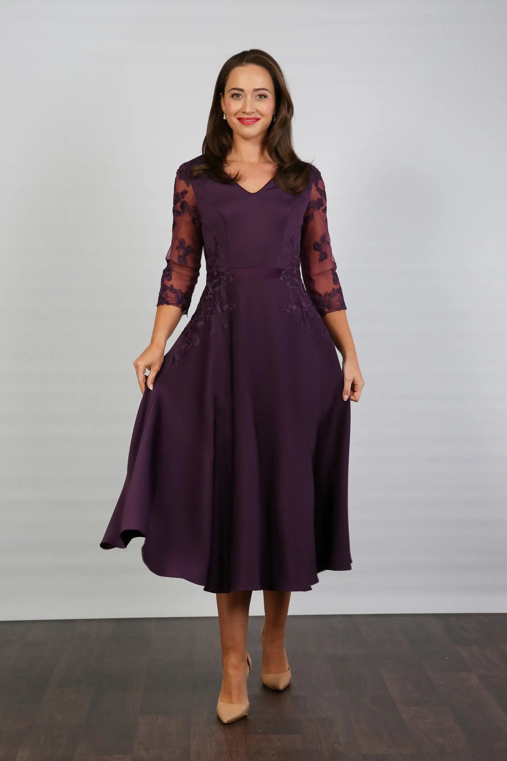 A woman models a midi length blackberry-coloured occasion wear dress with tulle sleeves and floral appliques.