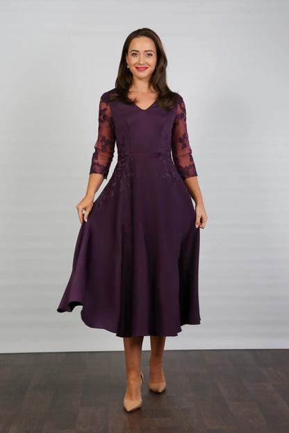 A woman models a midi length blackberry-coloured occasion wear dress with tulle sleeves and floral appliques.