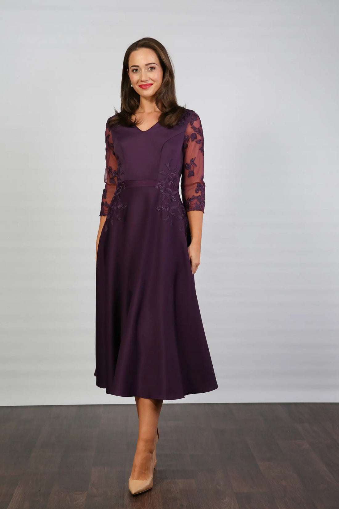 A woman models a midi length blackberry-coloured occasion wear dress with tulle sleeves and floral appliques.