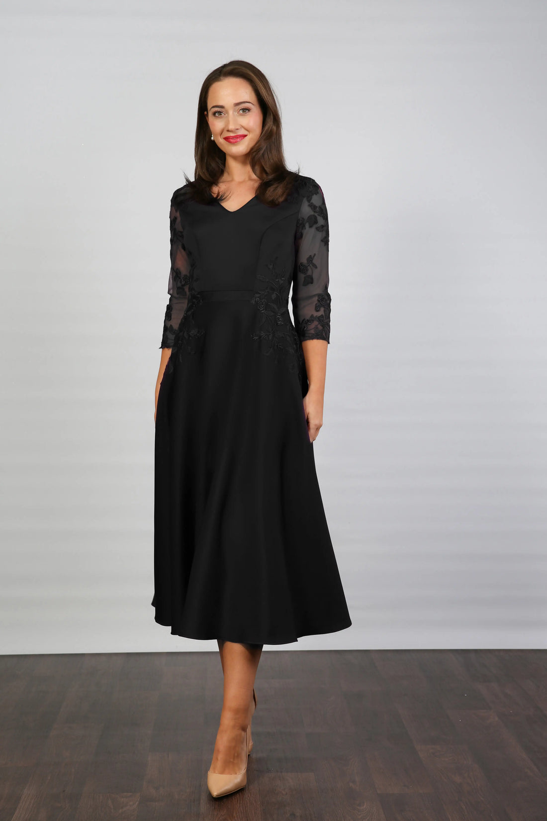 A woman models a midi length black occasion wear dress with tulle sleeves and floral appliques.