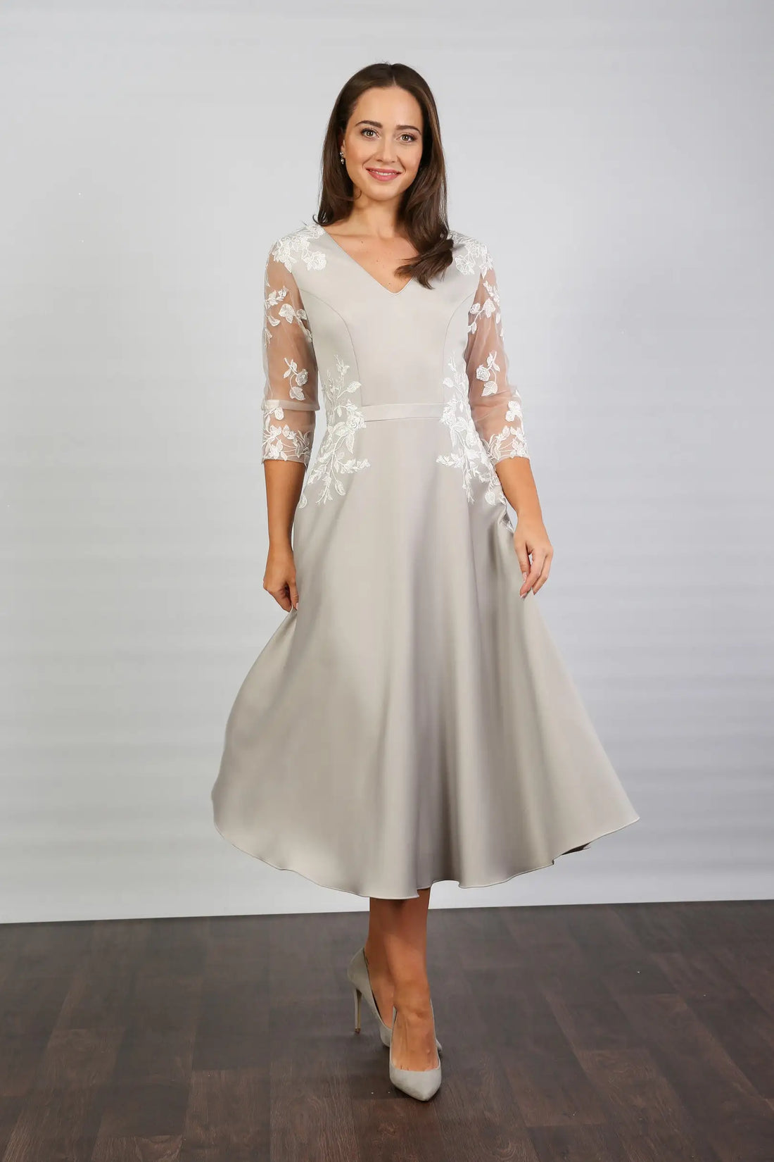 A woman modeling a midi length silver occasion dress with floral appliques on the sleeves and waist, featuring tulle sleeves and a v-neck.