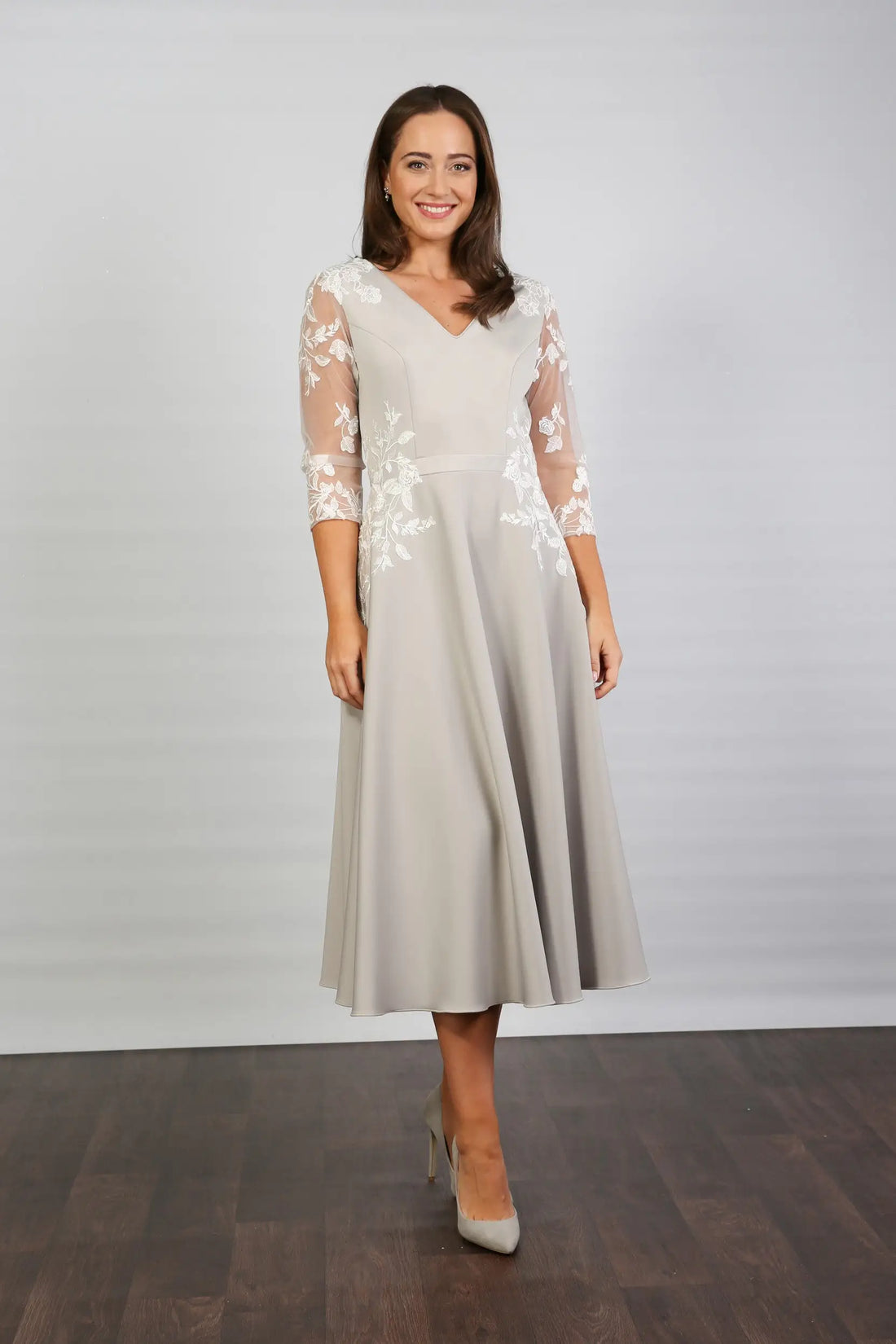 A woman modeling a midi length silver occasion dress with floral appliques on the sleeves and waist, featuring tulle sleeves and a v-neck.