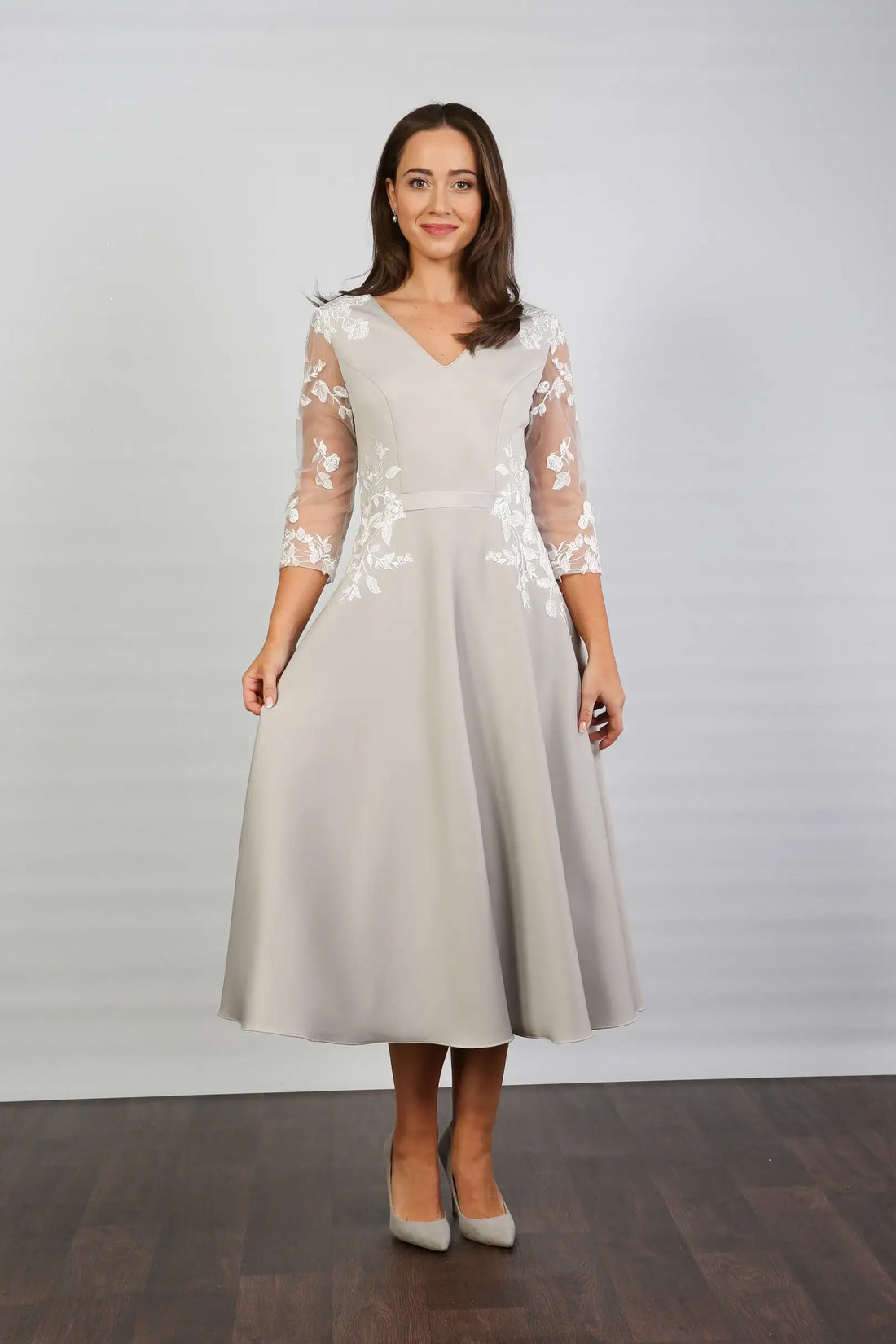 A woman modeling a midi length silver occasion dress with floral appliques on the sleeves and waist, featuring tulle sleeves and a v-neck.
