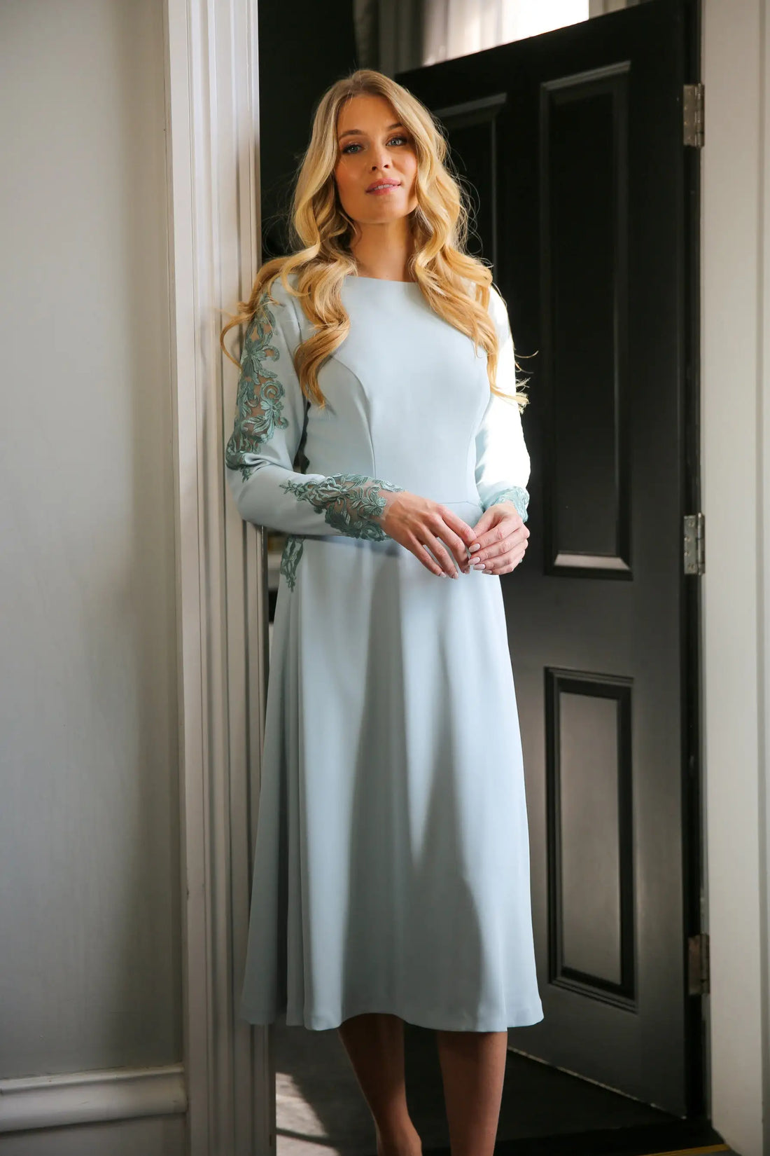 A woman wearing a long sleeve, midi length duck egg blue dress with embroidery detail at the waistline and sleeves.