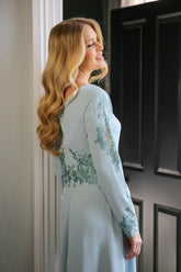 A woman wearing a long sleeve, midi length duck egg blue dress with embroidery detail at the waistline and sleeves.